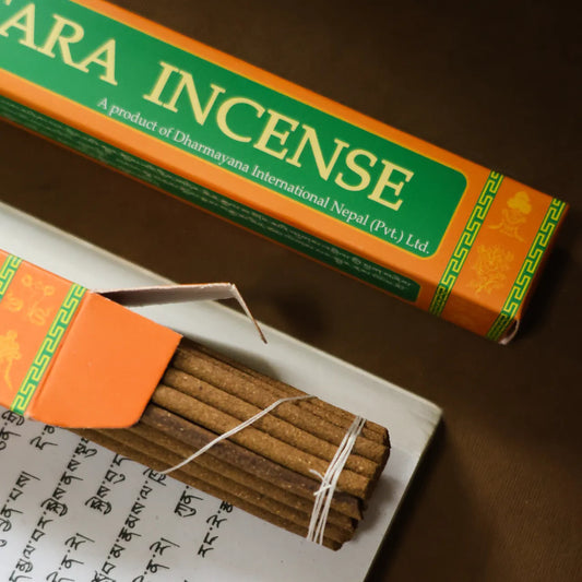 A bundle of incense sticks tied with white string, shown next to an open orange and green box labeled 'ARA INCENSE' by Dharmayana International, with a closed box placed above it on a dark brown surface.