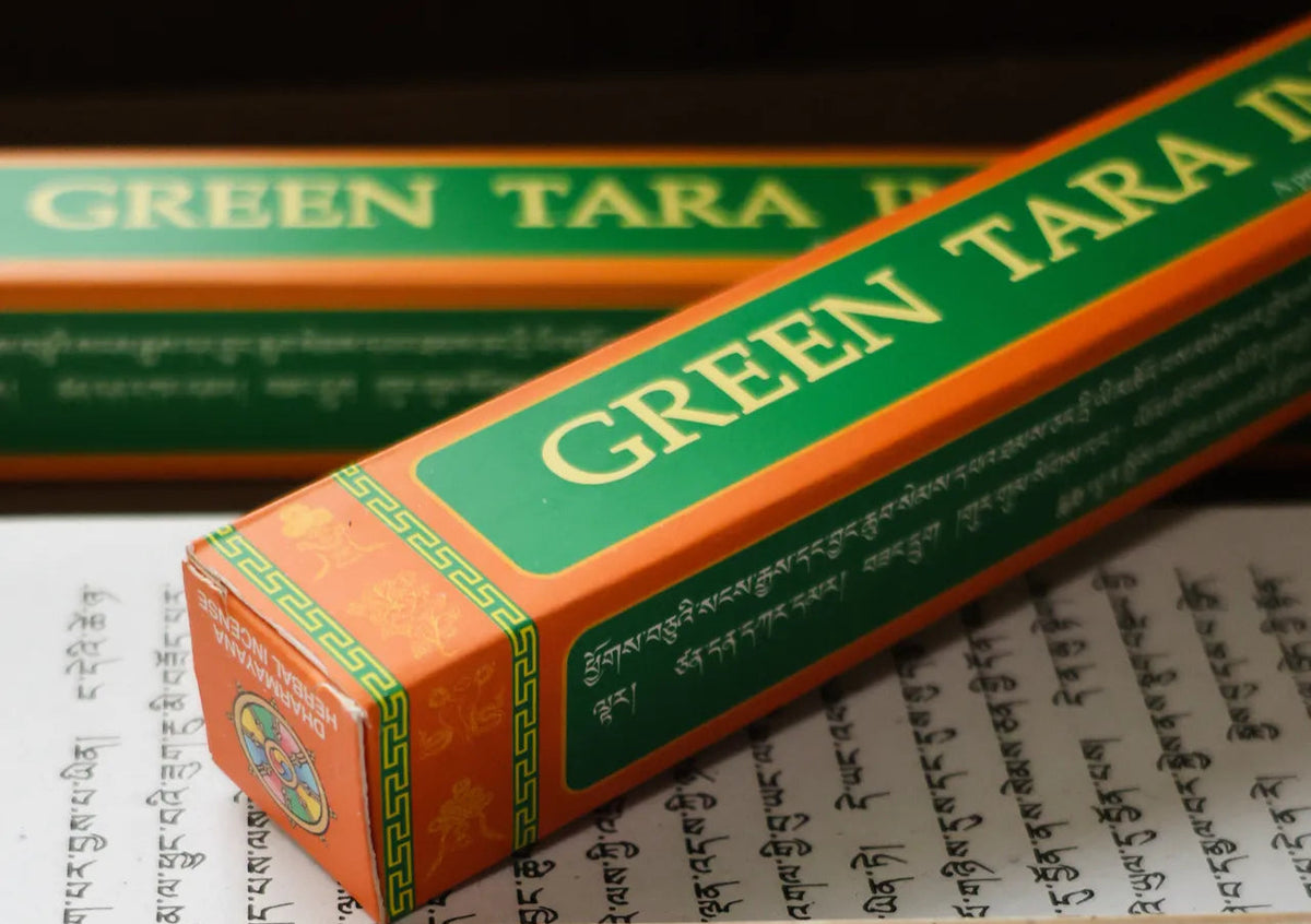 A close-up of a Green Tara incense stick box resting diagonally on top of an open book with printed text. The box is orange and green with gold lettering, and a single incense stick is placed in front of the box on a cream-colored card.